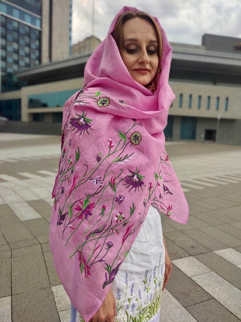 Puede incluir: Una bufanda de lino rosa con bordado floral. La bufanda est&aacute; drapeada sobre los hombros y la cabeza de una persona. El bordado es en tonos de morado, verde y blanco.