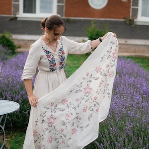 May include: A woman in a white linen dress with embroidered flowers stands in a field of lavender. She is holding a white linen scarf with a floral pattern.