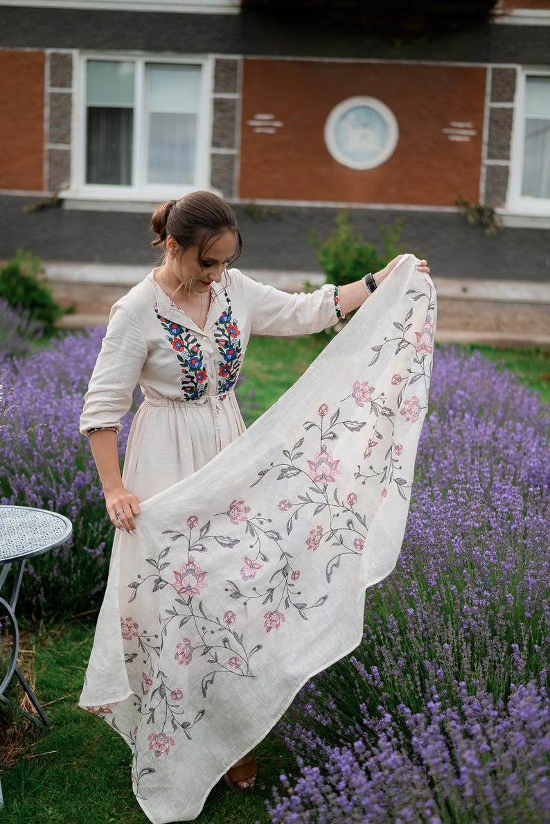 May include: A woman in a white linen dress with embroidered flowers stands in a field of lavender. She is holding a white linen scarf with a floral pattern.