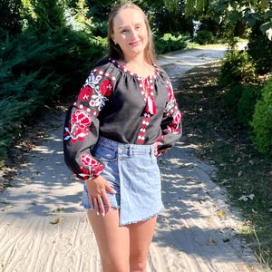 Puede incluir: Una mujer con una blusa negra bordada con dise&ntilde;os florales rojos y blancos, una falda de mezclilla azul claro y sandalias marrones. Est&aacute; de pie en un camino de arena.