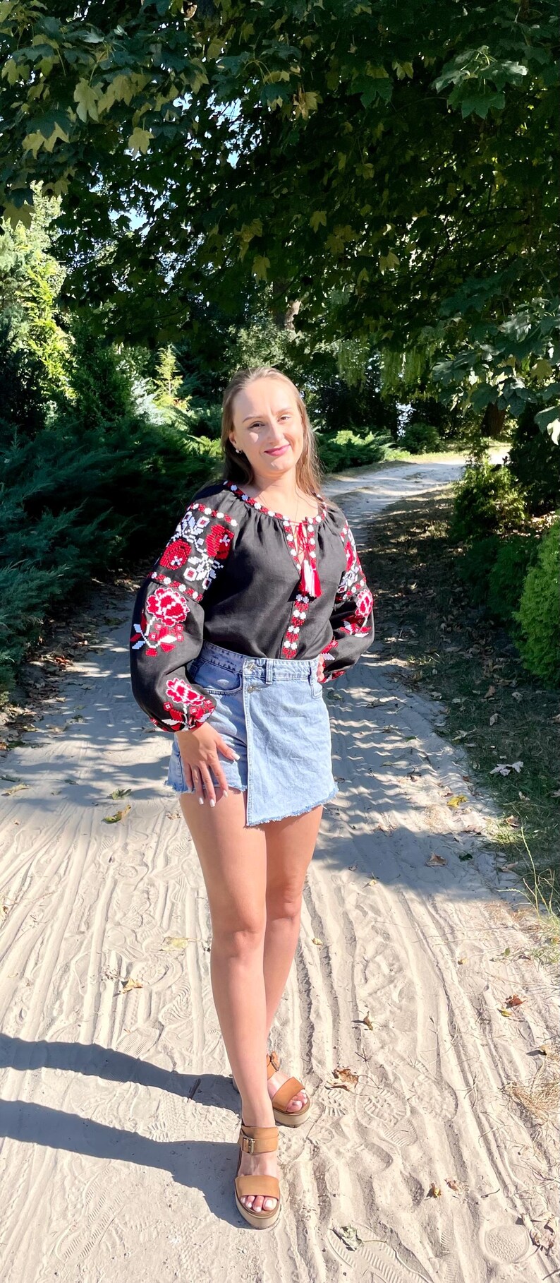Puede incluir: Una mujer con una blusa negra bordada con dise&ntilde;os florales rojos y blancos, una falda de mezclilla azul claro y sandalias marrones. Est&aacute; de pie en un camino de arena.