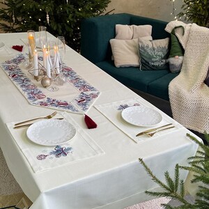Puede incluir: Una mesa navide&ntilde;a festiva con mantel blanco, camino de mesa bordado y manteles individuales. Cubiertos dorados y platos blancos est&aacute;n listos para una comida festiva. Los &aacute;rboles de Navidad y las luces decorativas crean un ambiente c&aacute;lido.