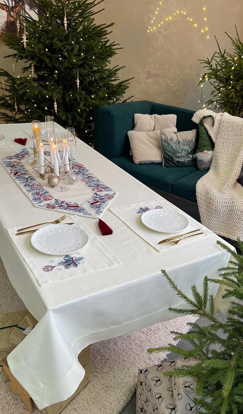 Puede incluir: Una mesa navide&ntilde;a festiva con mantel blanco, camino de mesa bordado y manteles individuales. Cubiertos dorados y platos blancos est&aacute;n listos para una comida festiva. Los &aacute;rboles de Navidad y las luces decorativas crean un ambiente c&aacute;lido.