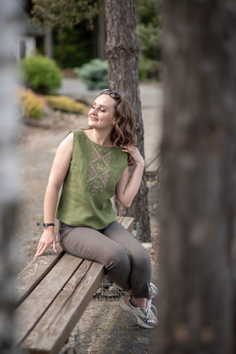 May include: A woman wearing a green sleeveless linen top with embroidered details sits on a wooden bench. She is wearing brown and white checked trousers and white trainers.