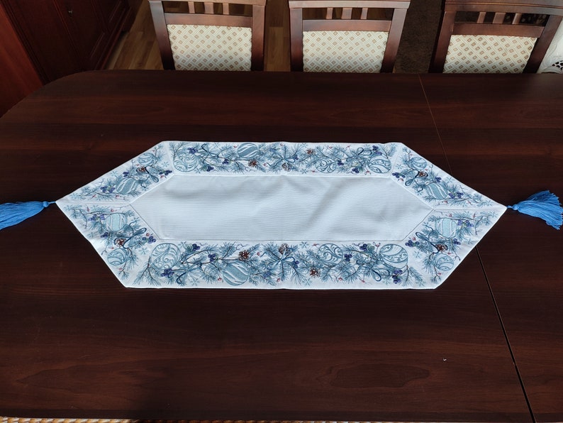 K&ouml;nnte beinhalten: Hellblau-wei&szlig;er Tischl&auml;ufer mit festlichem Design. Der L&auml;ufer hat eine hellblaue Mittelbahn und einen detaillierten Rand mit blauen Ornamenten, Tannennadeln und Beeren. Blaue Quasten schm&uuml;cken jedes Ende.