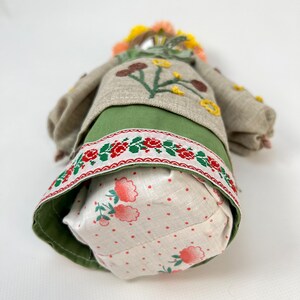 Puede incluir: Mu&ntilde;eca hecha a mano con una corona floral y detalles bordados. La mu&ntilde;eca lleva una falda verde, una blusa beige con aplicaciones de flores y una falda con un estampado floral rojo y blanco. El pelo de la mu&ntilde;eca es una mezcla de amarillo y naranja.