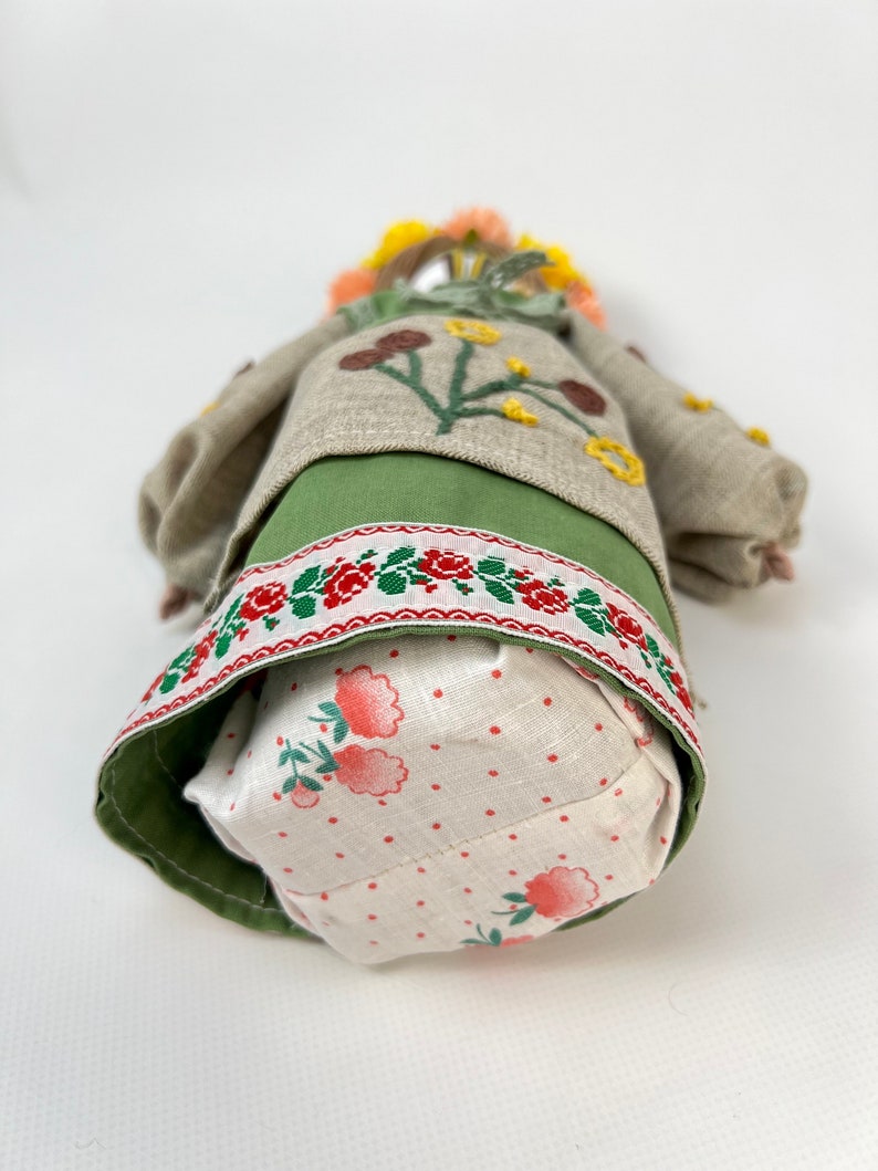 Puede incluir: Mu&ntilde;eca hecha a mano con una corona floral y detalles bordados. La mu&ntilde;eca lleva una falda verde, una blusa beige con aplicaciones de flores y una falda con un estampado floral rojo y blanco. El pelo de la mu&ntilde;eca es una mezcla de amarillo y naranja.