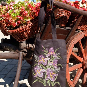 Op de afbeelding: Een zwarte tote bag met geborduurde paarse irissen. De tas hangt aan een houten kar met een rood wiel.