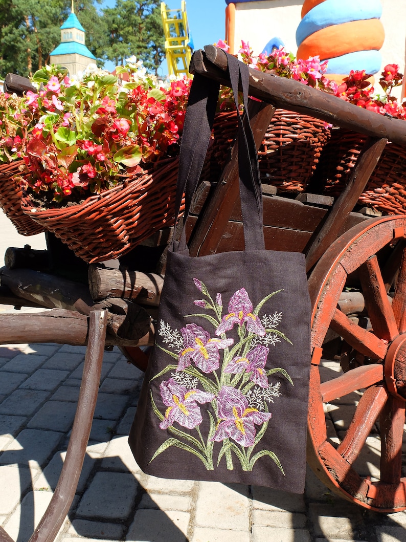 Op de afbeelding: Een zwarte tote bag met geborduurde paarse irissen. De tas hangt aan een houten kar met een rood wiel.