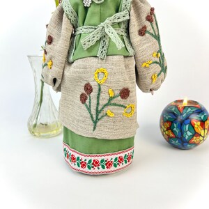 Puede incluir: Mu&ntilde;eca hecha a mano con un atuendo bordado verde y beige. El vestido presenta bordados florales amarillos y marrones. La falda tiene un ribete floral rojo y blanco. La mu&ntilde;eca lleva un collar de cuentas y un tocado floral.