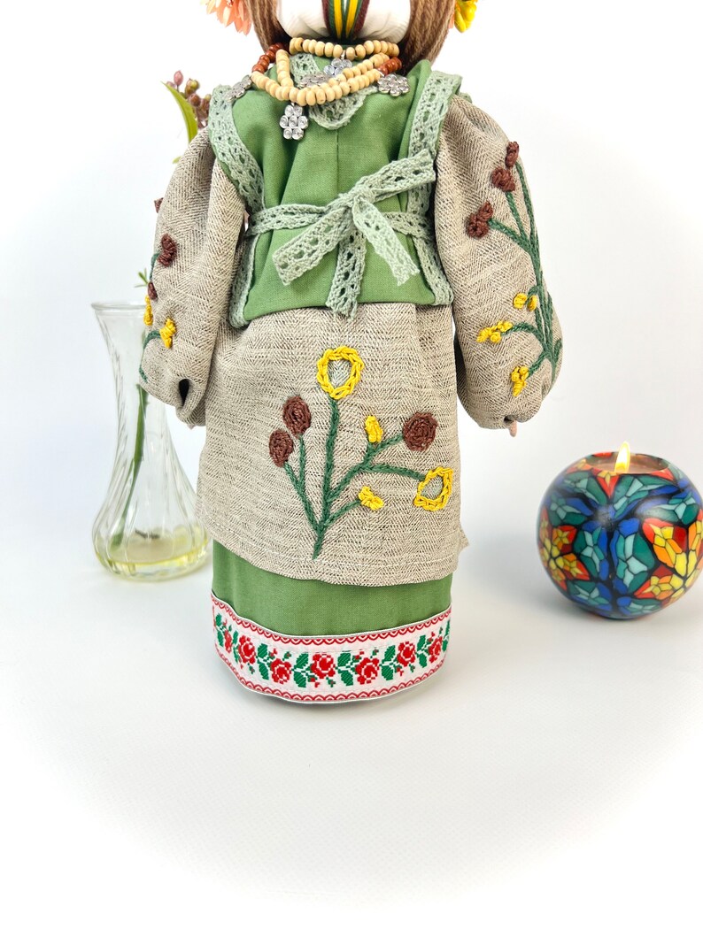 Puede incluir: Mu&ntilde;eca hecha a mano con un atuendo bordado verde y beige. El vestido presenta bordados florales amarillos y marrones. La falda tiene un ribete floral rojo y blanco. La mu&ntilde;eca lleva un collar de cuentas y un tocado floral.