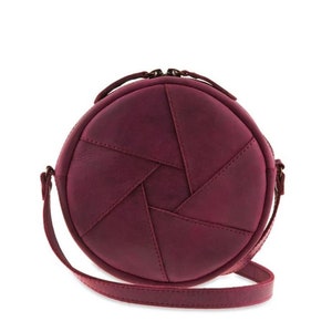 May include: A round, burgundy-colored crossbody bag with a unique design resembling a camera aperture. The bag features a stitched pattern and a long, thin strap. It has a top handle and a zipper closure.