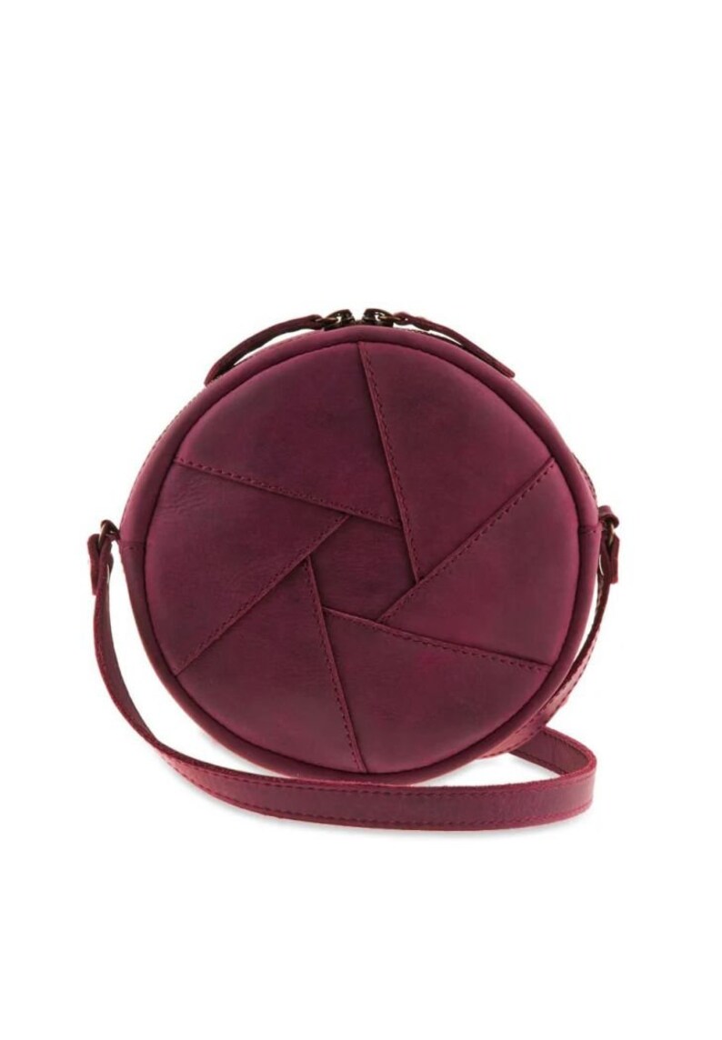 May include: A round, burgundy-colored crossbody bag with a unique design resembling a camera aperture. The bag features a stitched pattern and a long, thin strap. It has a top handle and a zipper closure.