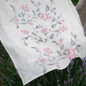 May include: White linen tablecloth with a delicate floral pattern in shades of pink and gray. The tablecloth is embroidered with a repeating design of flowers and leaves.