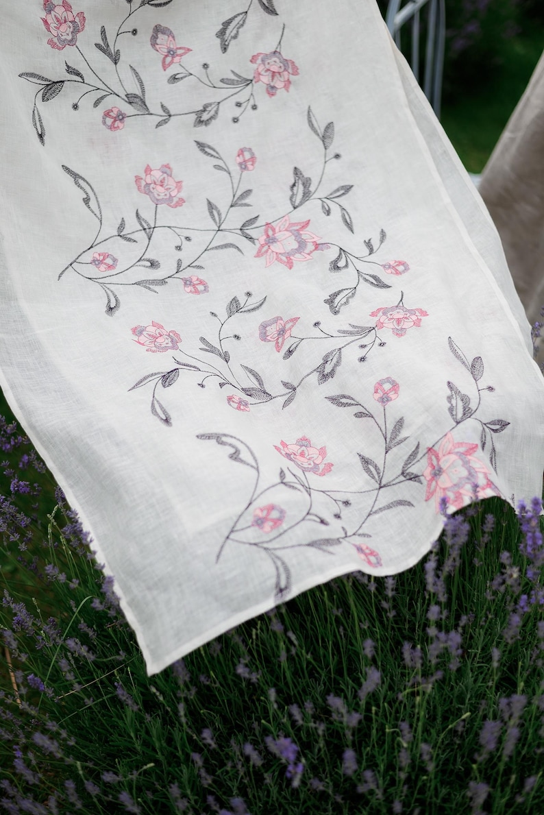 May include: White linen tablecloth with a delicate floral pattern in shades of pink and gray. The tablecloth is embroidered with a repeating design of flowers and leaves.
