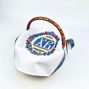 May include: A white cloth-lined basket with a woven handle. The cloth features a blue, yellow, and red embroidered design of an egg shape with text. The rim of the cloth has a blue patterned border.