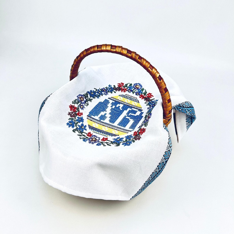 May include: A white cloth-lined basket with a woven handle. The cloth features a blue, yellow, and red embroidered design of an egg shape with text. The rim of the cloth has a blue patterned border.