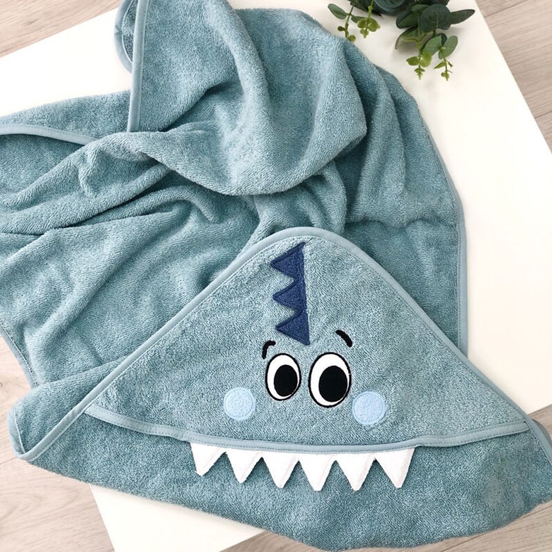May include: A teal hooded towel with a cute embroidered dinosaur face. The dinosaur has big eyes, a toothy grin, and a spiky crest.