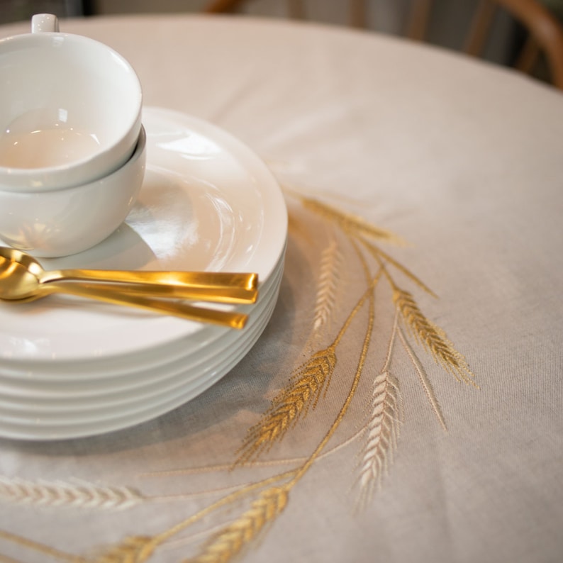 Peut inclure: Une pile d'assiettes en c&eacute;ramique blanche et une tasse et une soucoupe blanches reposent sur une nappe beige. Deux cuill&egrave;res dor&eacute;es sont plac&eacute;es &agrave; c&ocirc;t&eacute; des assiettes. La nappe pr&eacute;sente un motif de bl&eacute; brod&eacute;.
