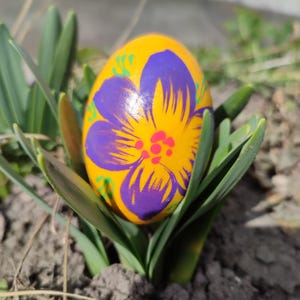 May include: A hand-painted Easter egg with a vibrant yellow base and a large purple flower design. The flower's center is red, and green accents surround the petals. The egg is nestled among green plant leaves, suggesting a spring theme.