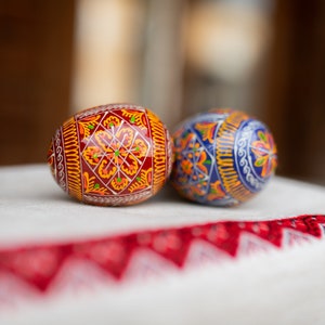 K&ouml;nnte beinhalten: Zwei verzierte Ostereier mit aufwendigen Mustern. Ein Ei ist haupts&auml;chlich rot mit gelben und orangenen Designs, das andere blau mit orangefarbenen und silbernen Akzenten. Sie liegen auf einem wei&szlig;en Tuch mit roter Stickerei, das traditionelle Handwerkskunst zeigt.