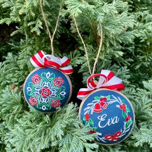 K&ouml;nnte beinhalten: Zwei blaue Weihnachtskugeln mit wei&szlig;en und roten Stickereien. Eine Kugel hat ein Blumenmuster und die andere einen Kranz mit Glocken und dem Namen "Eva" bestickt.