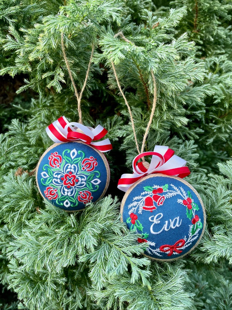K&ouml;nnte beinhalten: Zwei blaue Weihnachtskugeln mit wei&szlig;en und roten Stickereien. Eine Kugel hat ein Blumenmuster und die andere einen Kranz mit Glocken und dem Namen "Eva" bestickt.