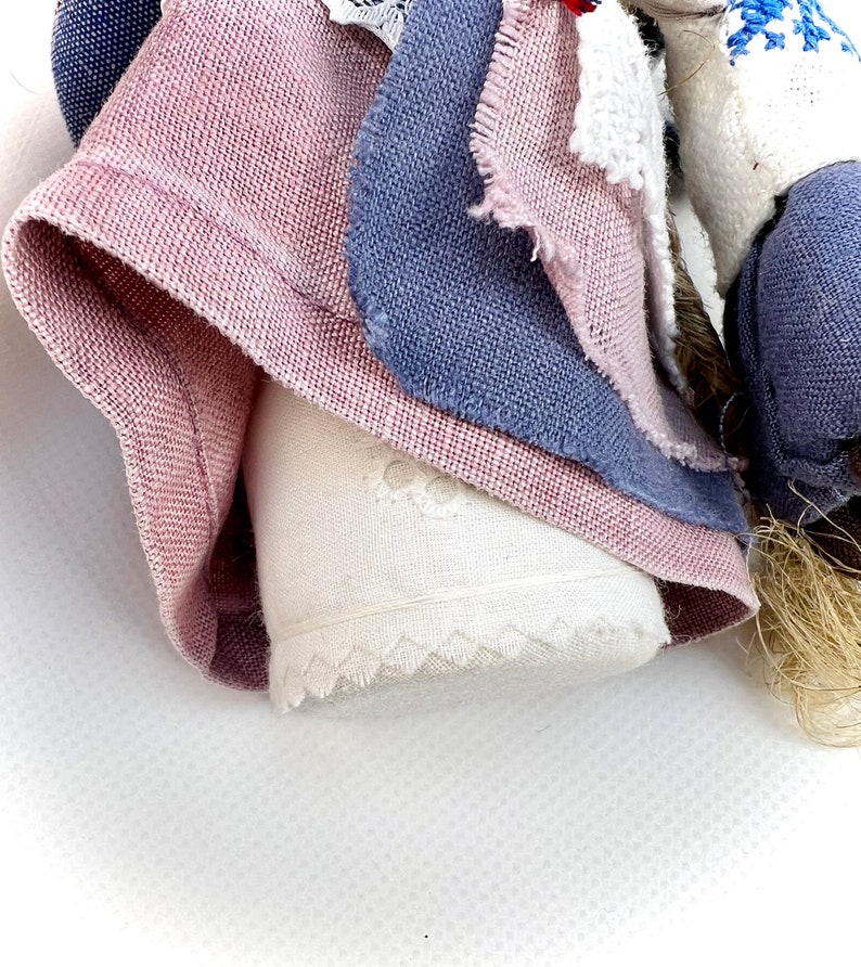 May include: A handmade rag doll with a pink, blue, and white fabric dress. The doll has a white fabric face with embroidered details.