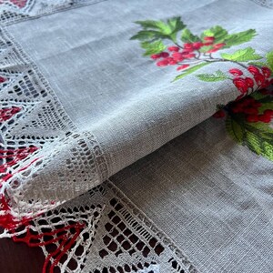 May include: A gray linen table runner with a red and white lace border. The runner features an embroidered design of a green holly sprig with red berries.