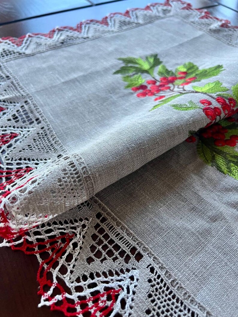 May include: A gray linen table runner with a red and white lace border. The runner features an embroidered design of a green holly sprig with red berries.