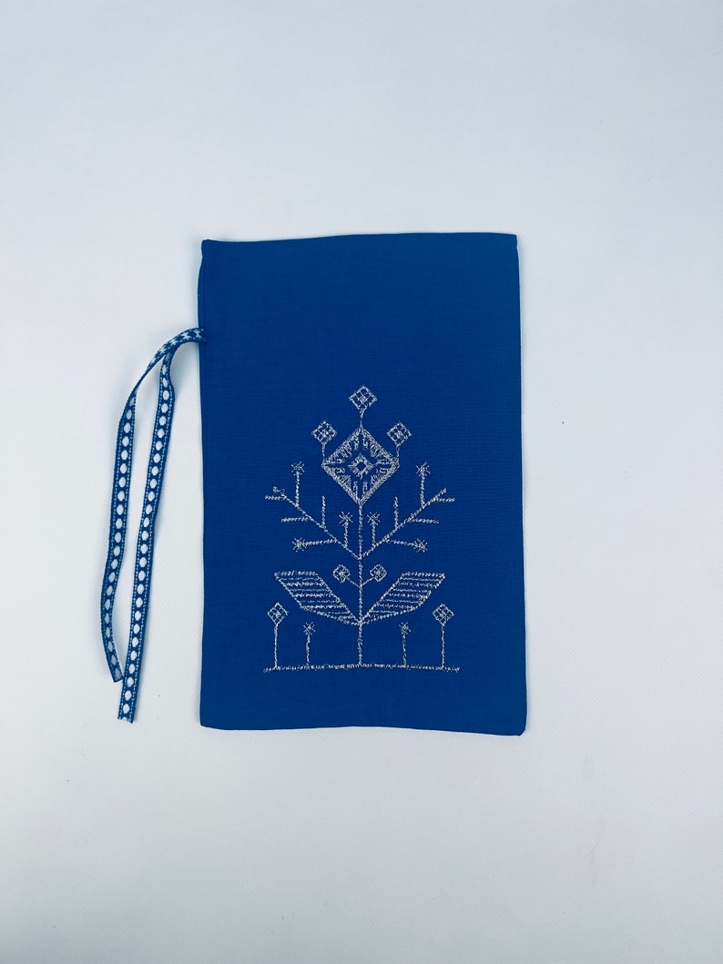 K&ouml;nnte beinhalten: Eine blaue Stofftasche mit einem wei&szlig;en gestickten Design einer stilisierten Blume. Die Tasche hat einen wei&szlig;en und blauen Kordelzug.
