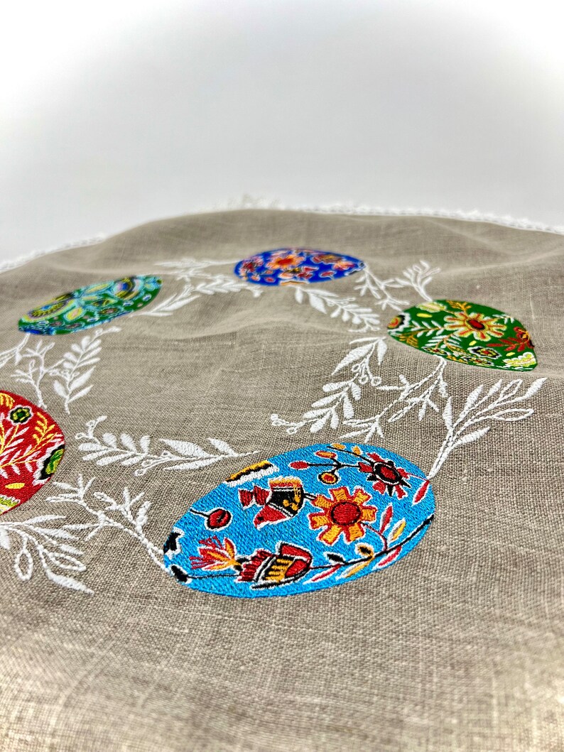 以下が含まれることがあります： ベージュのリネン製のテーブルランナーに、白い刺繍の枝とカラフルなイースターエッグが施されています。卵は、青、赤、緑、黄色の花柄で飾られています。