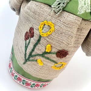 Puede incluir: Mu&ntilde;eca de tela con detalles florales bordados. La mu&ntilde;eca presenta un cuerpo de tela beige con estampado de espiga, detalles verdes y una cinta floral decorativa. Flores bordadas amarillas y marrones adornan el frente.