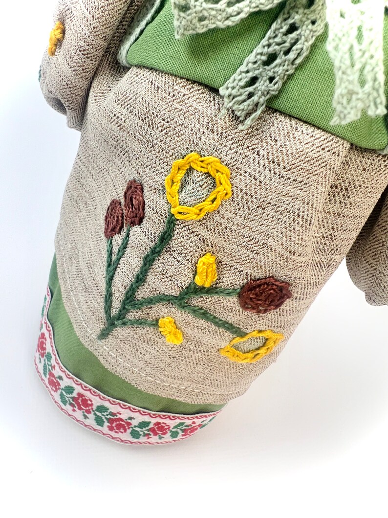 Puede incluir: Mu&ntilde;eca de tela con detalles florales bordados. La mu&ntilde;eca presenta un cuerpo de tela beige con estampado de espiga, detalles verdes y una cinta floral decorativa. Flores bordadas amarillas y marrones adornan el frente.