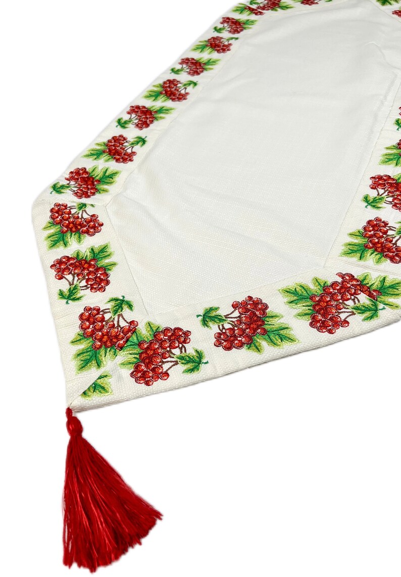Puede incluir: Camino de mesa blanco con un borde detallado con bayas rojas y hojas verdes. Una borla roja cuelga de la esquina. El camino de mesa est&aacute; hecho de una tela texturizada, adecuada para la decoraci&oacute;n del hogar.