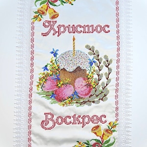 May include: Embroidered white fabric featuring the words "Christ is Risen" in red script. The design includes a decorated cake with a lit candle, Easter eggs, flowers, and bells, framed by a lace border.