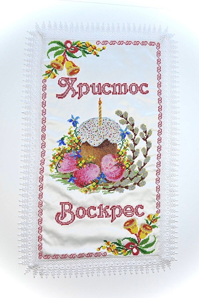 May include: Embroidered white fabric featuring the words "Christ is Risen" in red script. The design includes a decorated cake with a lit candle, Easter eggs, flowers, and bells, framed by a lace border.