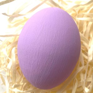 May include: A large, lavender egg with a textured surface, set on a bed of light-coloured, shredded paper. The egg's surface has visible brushstrokes, giving it a handcrafted appearance. The overall theme suggests Easter or springtime decor.