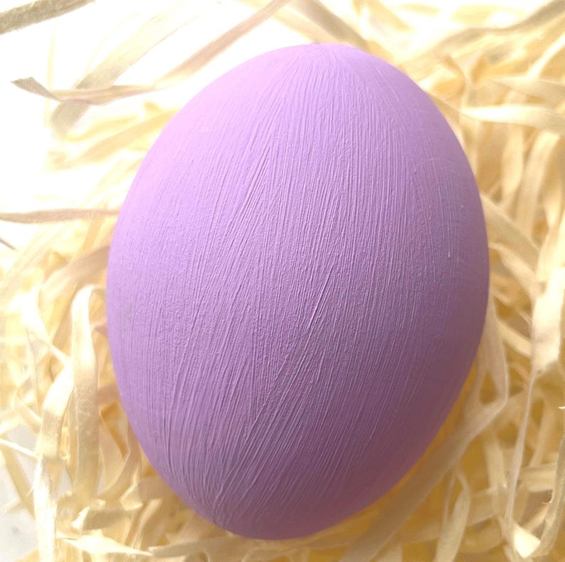 May include: A large, lavender egg with a textured surface, set on a bed of light-coloured, shredded paper. The egg's surface has visible brushstrokes, giving it a handcrafted appearance. The overall theme suggests Easter or springtime decor.
