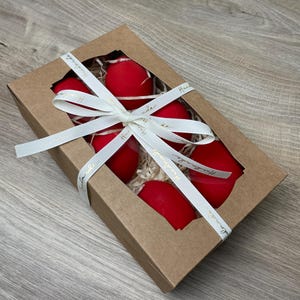 May include: A gift box with a clear window contains six bright red, egg-shaped objects. The box is tied with a white ribbon with gold text. The box is made of brown cardboard and filled with straw.