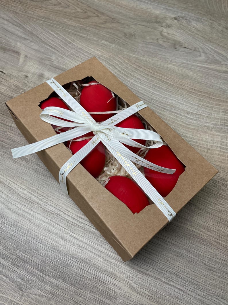 May include: A gift box with a clear window contains six bright red, egg-shaped objects. The box is tied with a white ribbon with gold text. The box is made of brown cardboard and filled with straw.