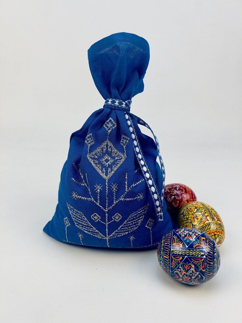 Pode incluir: Uma bolsa azul royal com cord&atilde;o e bordado prateado, fita branca e azul. Tr&ecirc;s ovos de P&aacute;scoa decorados em vermelho, amarelo e azul est&atilde;o ao lado da bolsa. A bolsa tem um design floral.