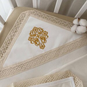 May include: White crib sheet with a gold embroidered cross design and a lace trim. The sheet is on a white crib mattress.