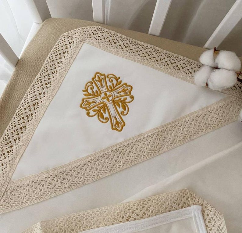 May include: White crib sheet with a gold embroidered cross design and a lace trim. The sheet is on a white crib mattress.