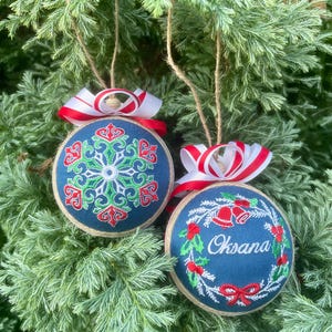 K&ouml;nnte beinhalten: Zwei handgemachte Weihnachtsbaumschmuck mit rot-wei&szlig;em Band. Ein Ornament zeigt ein gr&uuml;nes und rotes Schneeflockenmuster auf blauem Hintergrund. Das andere Ornament zeigt einen gr&uuml;nen Kranz mit roten Glocken und dem Namen "Oksana" auf blauem Hintergrund.