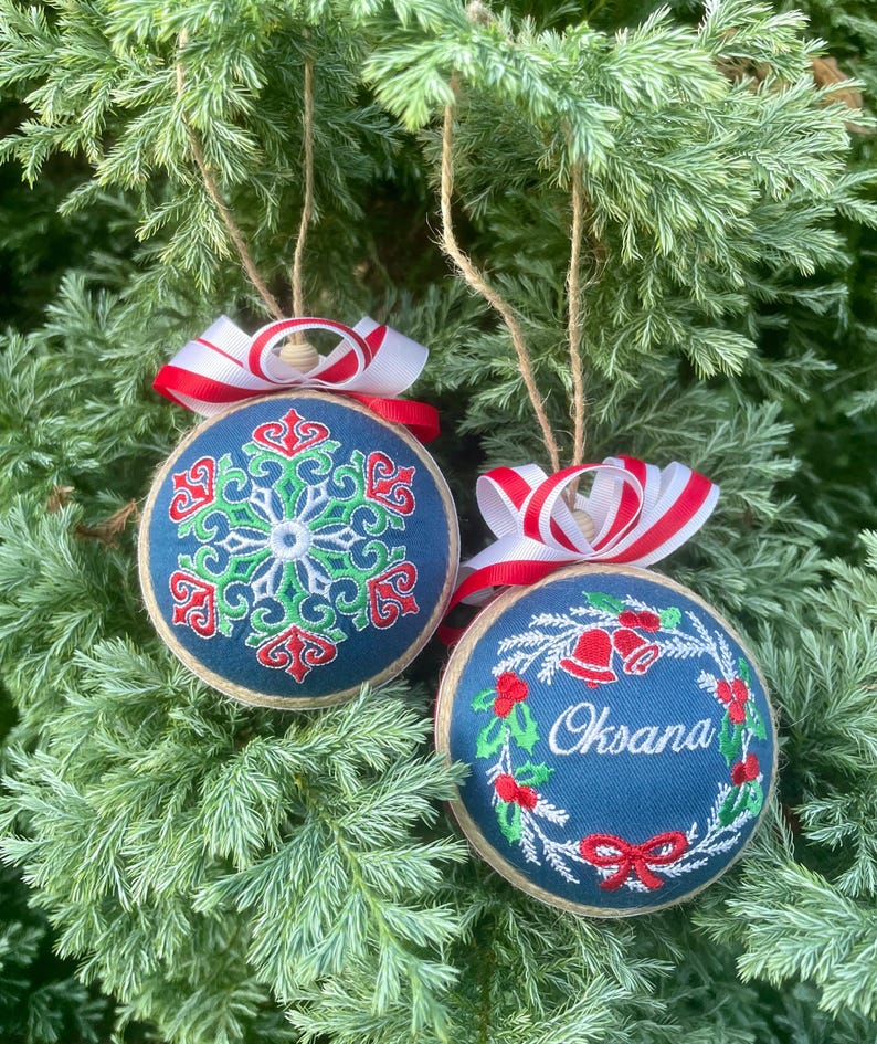K&ouml;nnte beinhalten: Zwei handgemachte Weihnachtsbaumschmuck mit rot-wei&szlig;em Band. Ein Ornament zeigt ein gr&uuml;nes und rotes Schneeflockenmuster auf blauem Hintergrund. Das andere Ornament zeigt einen gr&uuml;nen Kranz mit roten Glocken und dem Namen "Oksana" auf blauem Hintergrund.