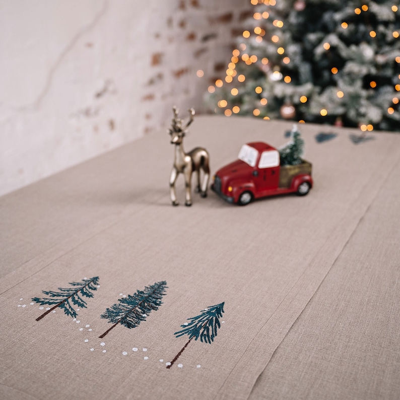 May include: A beige linen table runner with embroidered Christmas trees and white accents. A red toy truck with a Christmas tree in the bed and a gold reindeer figurine are on the runner. A Christmas tree is in the background.