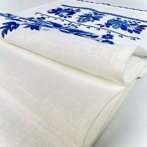 Peut inclure: Chemin de table en lin blanc avec broderie florale bleue. Le chemin de table est pli&eacute;, r&eacute;v&eacute;lant la texture du tissu et les motifs de broderie d&eacute;taill&eacute;s. Une pi&egrave;ce d&eacute;corative pour une table.