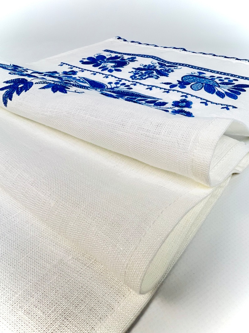 Peut inclure: Chemin de table en lin blanc avec broderie florale bleue. Le chemin de table est pli&eacute;, r&eacute;v&eacute;lant la texture du tissu et les motifs de broderie d&eacute;taill&eacute;s. Une pi&egrave;ce d&eacute;corative pour une table.