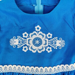 K&ouml;nnte beinhalten: Ein blaues Leinenkleid mit wei&szlig;en gestickten Blumenmotiven und einem wei&szlig;en Spitzenbesatz am Saum.